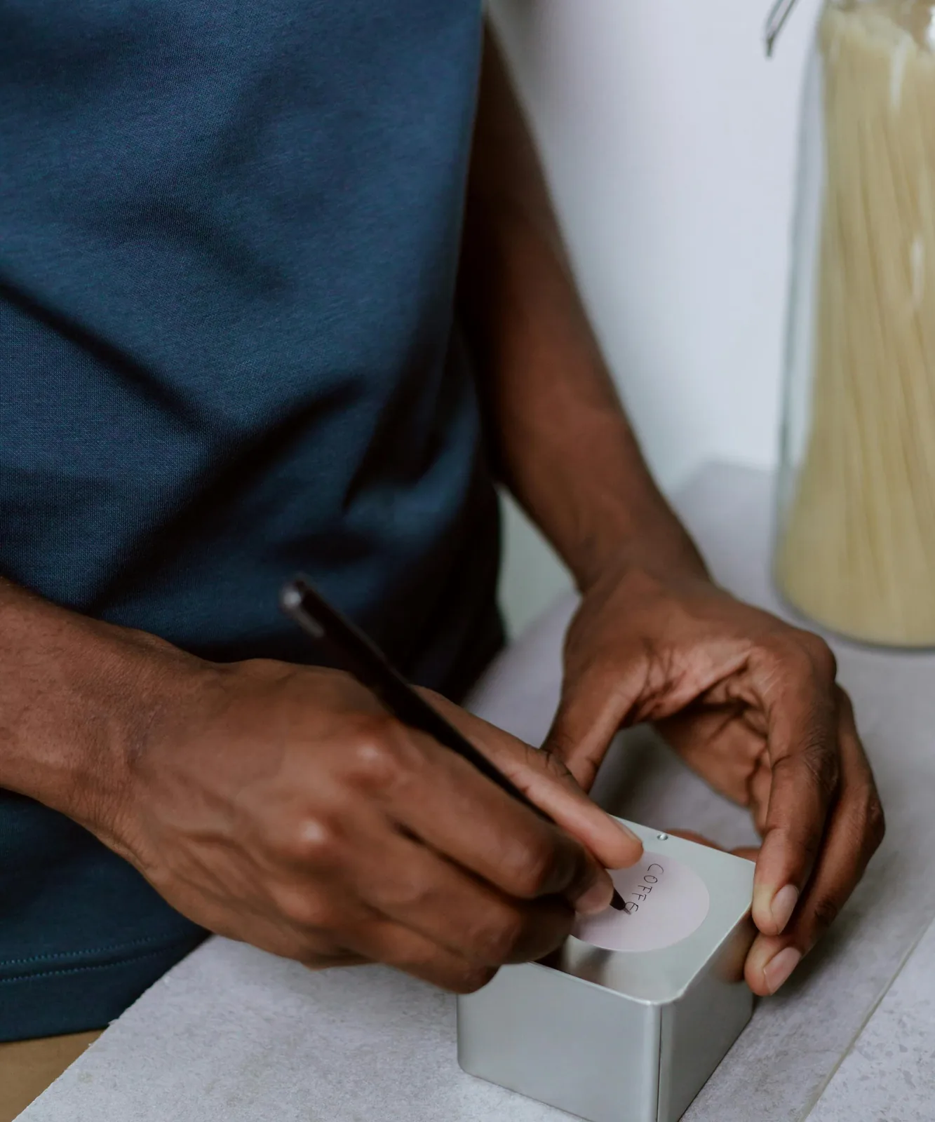 Hands labeling a small tin at a kitchen workbench with a glass jar of pasta