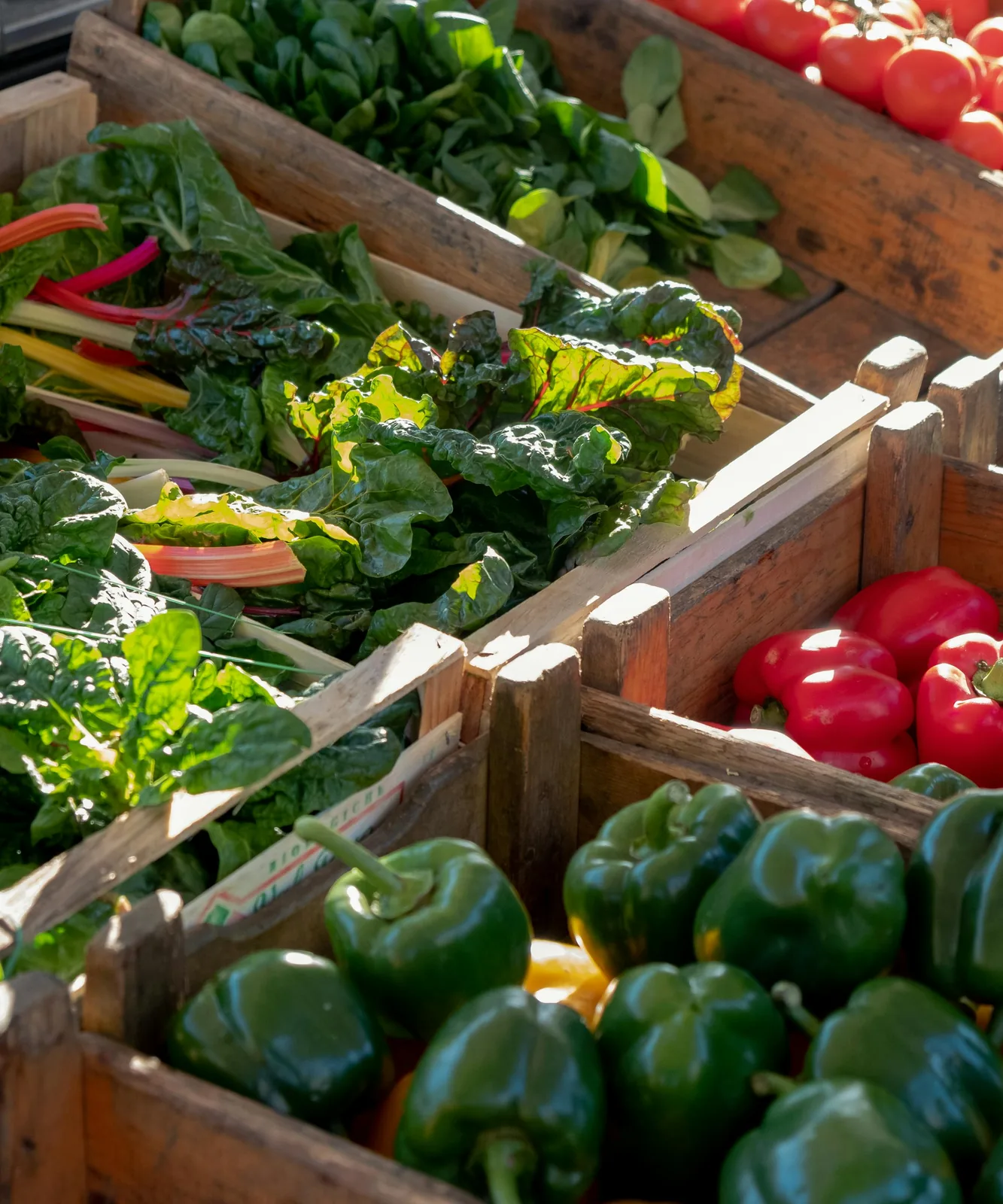 Wooden market crates filled with peppers, tomatoes, and rainbow chard in sunlight