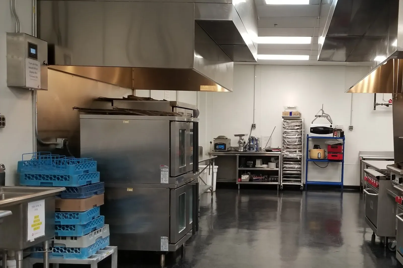 Commercial kitchen with double convection ovens, stainless steel prep stations, industrial exhaust hoods, and polished concrete floors